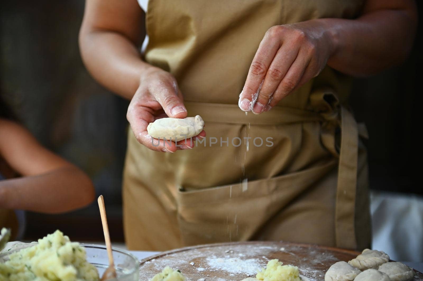 Details: hands of a housewife in chef's apron, sprinkling white flour on a wooden board while hands-on dumplings, cooking homemade varenyky in the country kitchen. Scene of artisanal food preparation