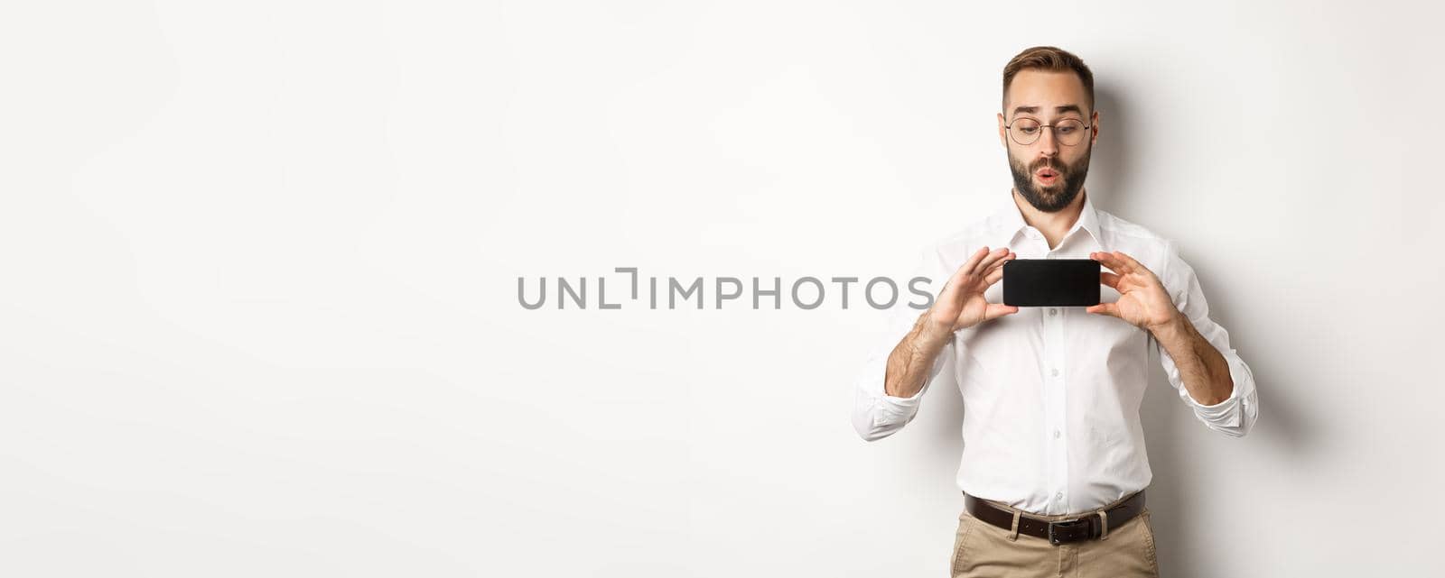 Amused handsome guy showing mobile screen, looking excited at online website, standing over white background.