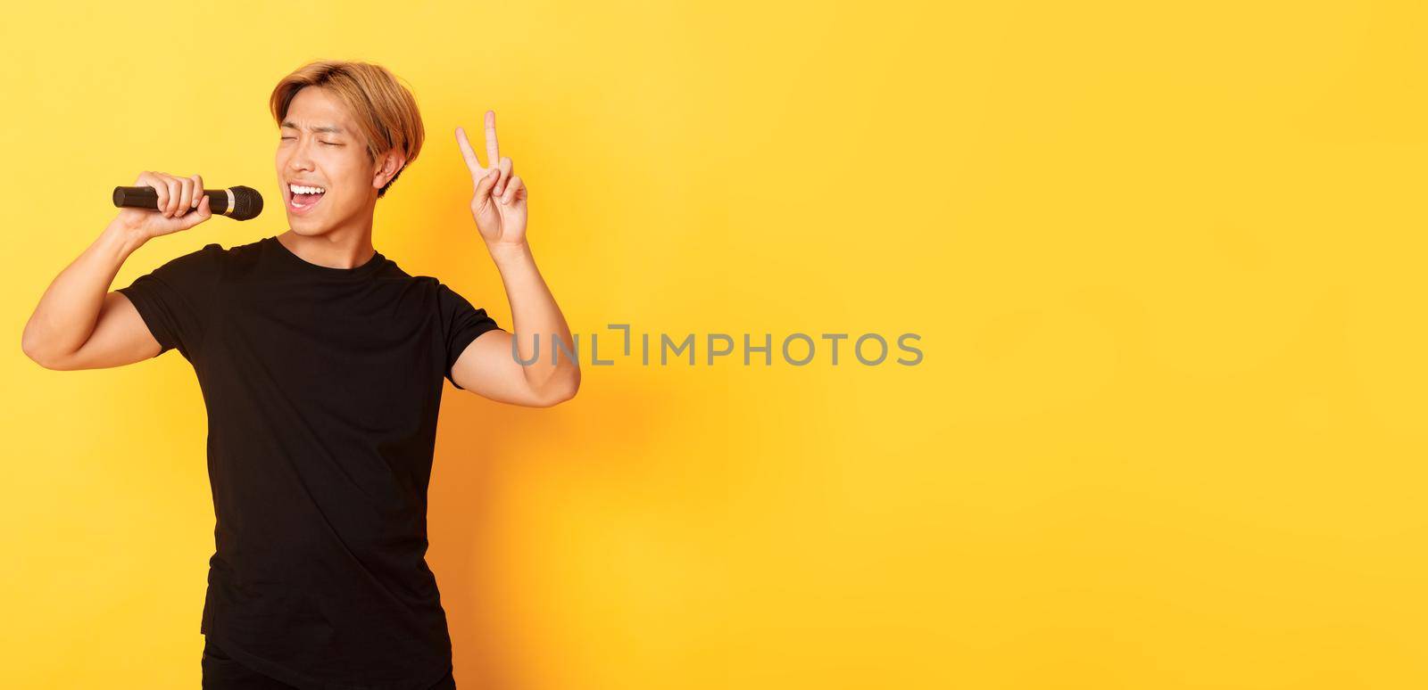 Carefree handsome asian guy performing song, singing into microphone and showing peace gesture, standing yellow background by Benzoix