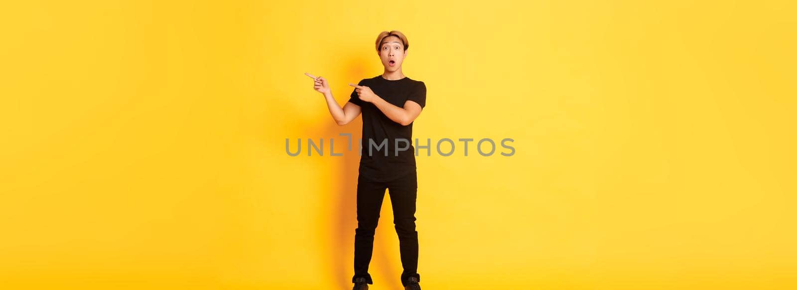Full-length of amazed and impressed attractive asian guy pointing fingers upper right corner, showing logo, yellow background by Benzoix