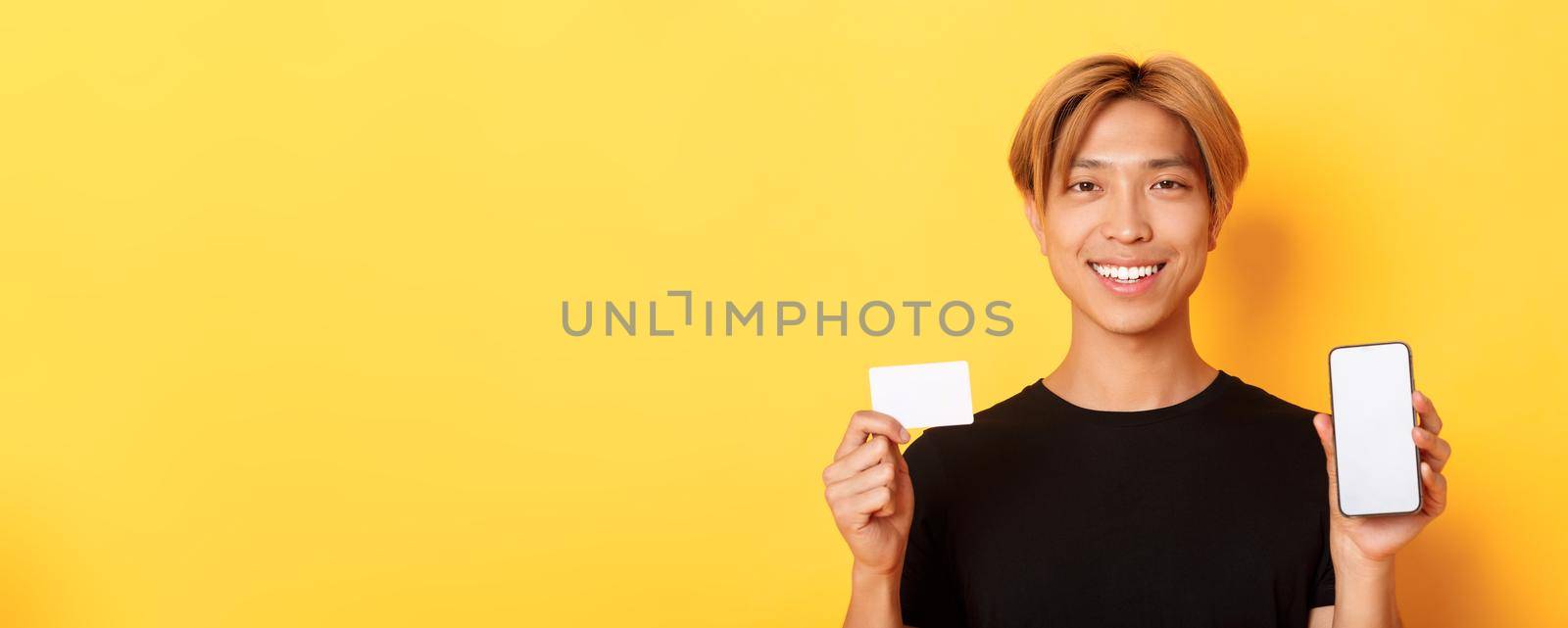 Close-up of smiling handsome asian guy showing smartphone screen and credit card, standing yellow background by Benzoix