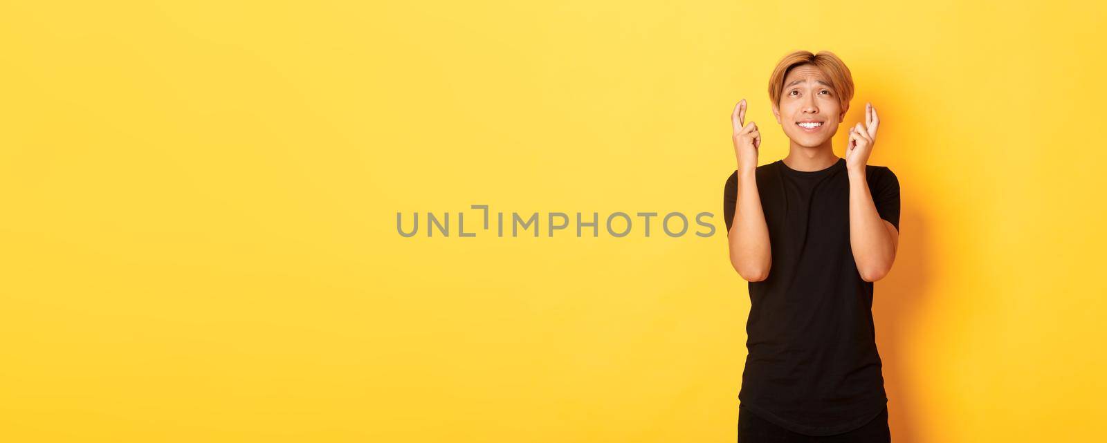 Portrait of hopeful worried asian man cross fingers good luck, begging look up, standing yellow background by Benzoix