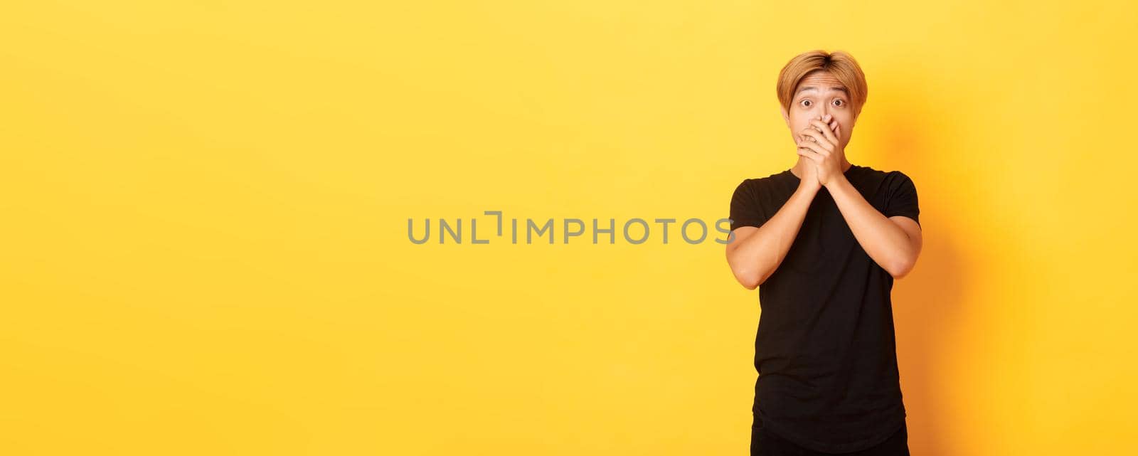 Portrait of surprised gasping asian guy with blond haircut, cover mouth with hands and looking amazed, standing yellow background.