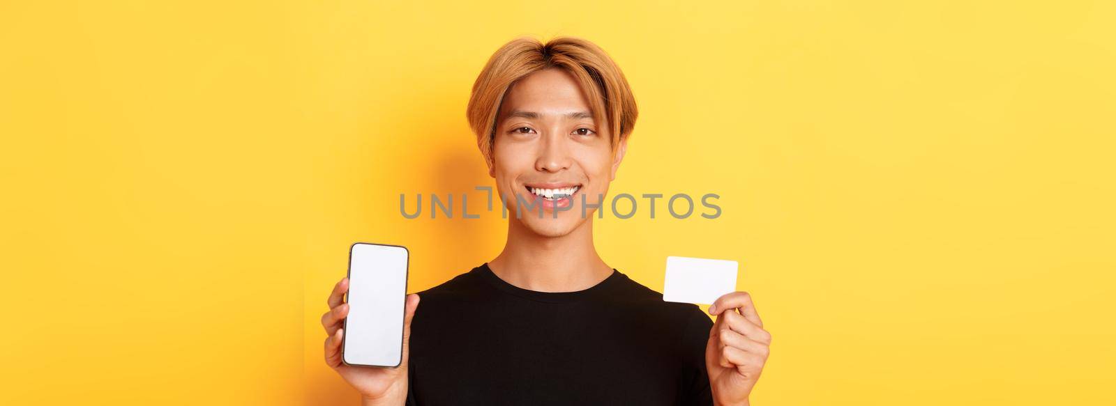 Close-up of smiling handsome asian guy showing smartphone screen and credit card, standing yellow background.