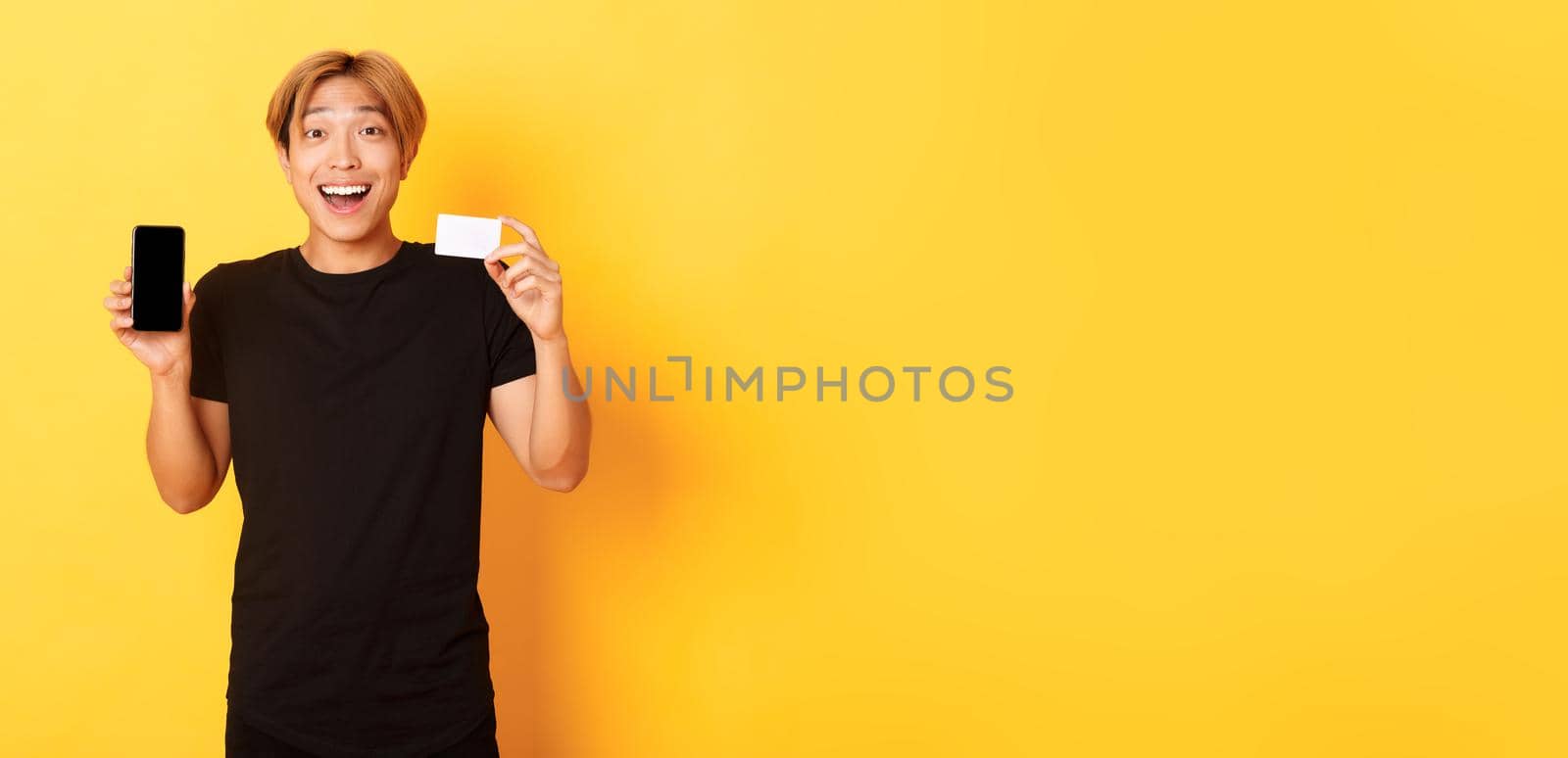 Portrait of excited happy asian man showing mobile phone screen and credit card with joyful smile, standing yellow background.