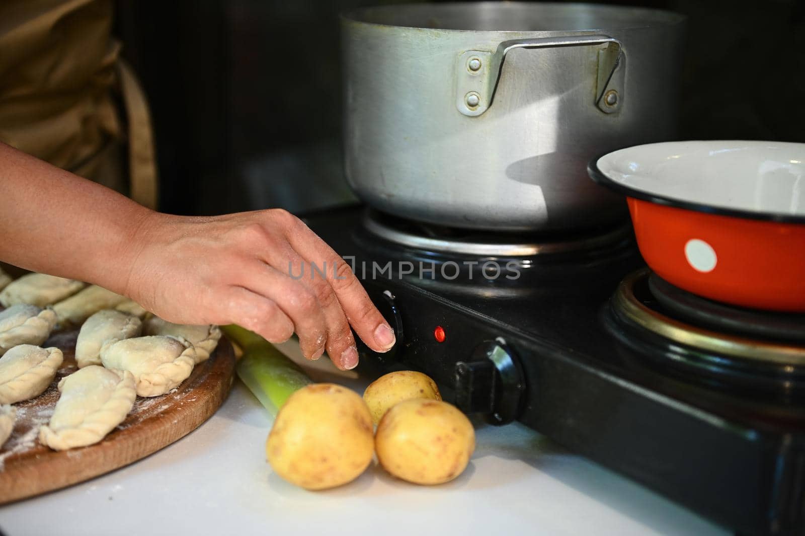 Close-up of a housewife switching on the electric stove, while preparing homemade dumplings for dinner. Unrecognizable woman chef cooking Ukrainian varenyky in the country kitchen. Pelmeni Ravioli
