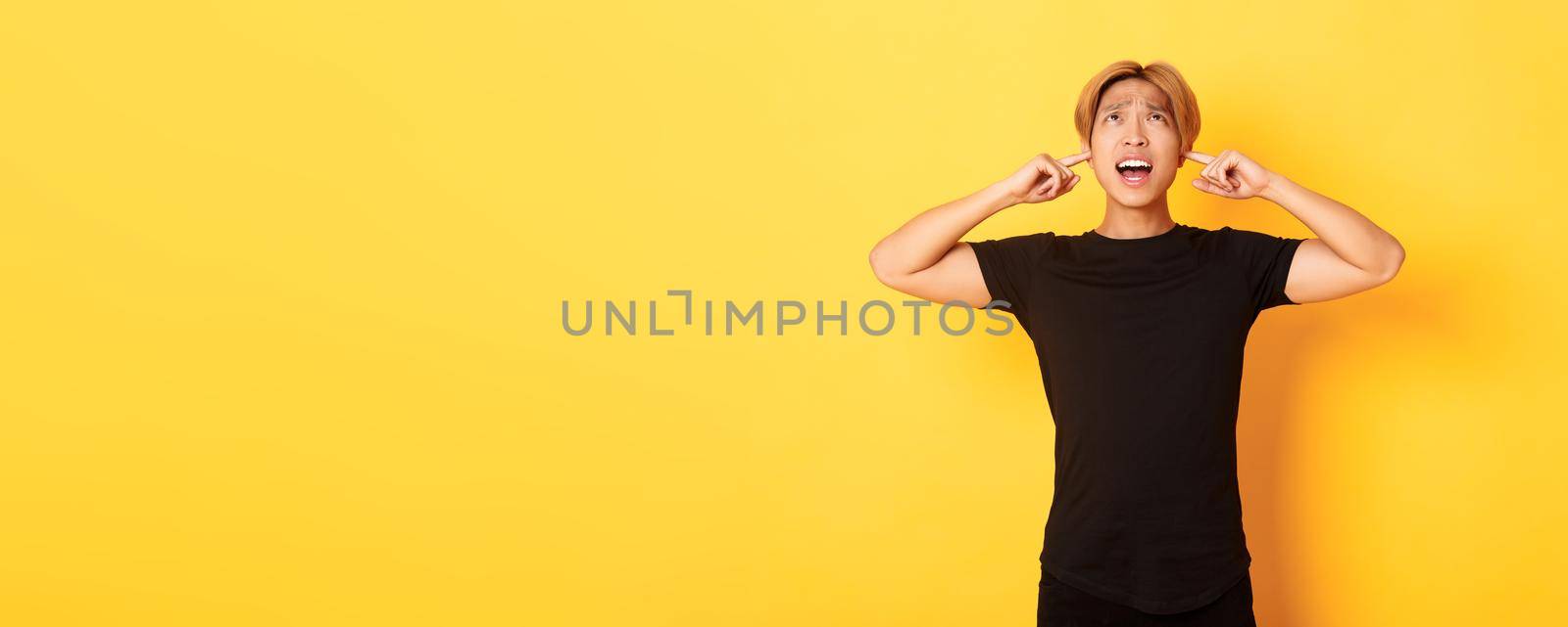 Portrait of annoyed and bothered asian guy grimacing, shut ears disturbed by loud noise, standing yellow background.