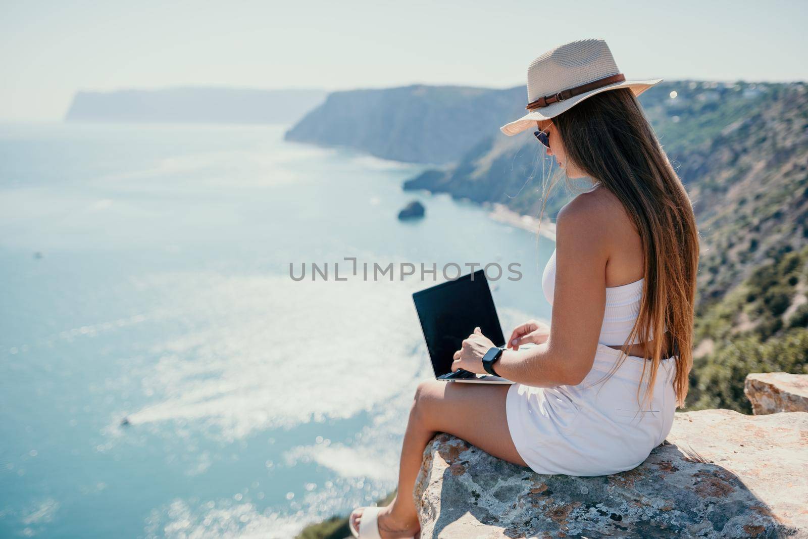 Successful business woman in yellow hat working on laptop by the sea. Pretty lady typing on computer at summer day outdoors. Freelance, travel and holidays concept.