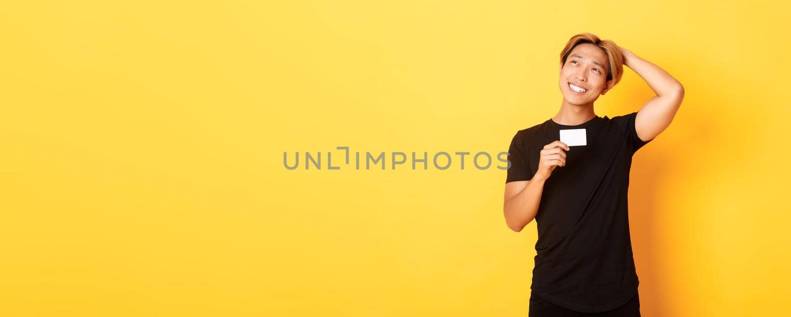 Thoughtful smiling asian guy thinking while showing credit card, looking upper left corner dreamy, yellow background by Benzoix