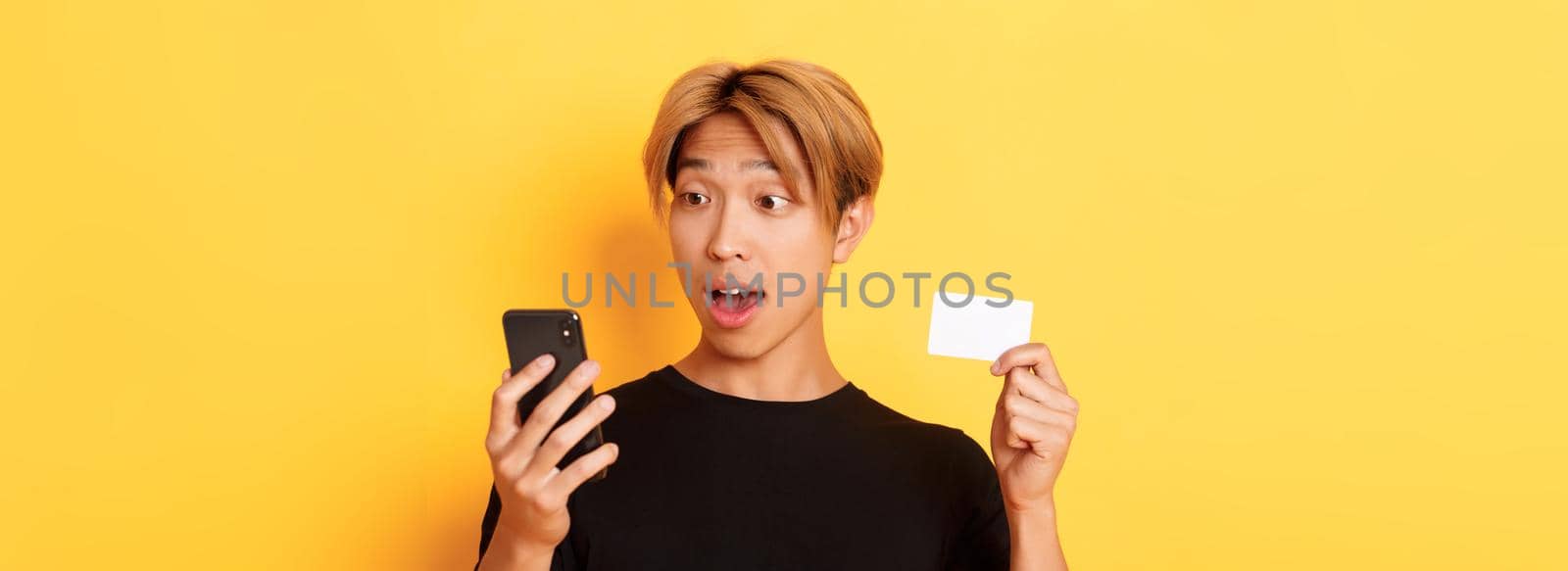 Close-up of amazed asian guy looking at mobile phone while shopping online and showing credit card, yellow background by Benzoix