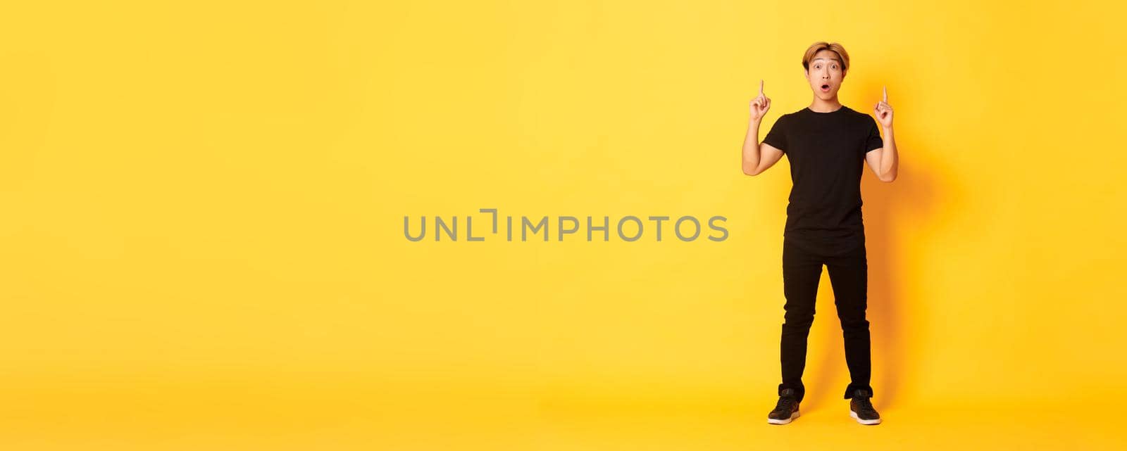 Full-length of impressed asian guy in black clothes, gasping amazed and pointing fingers up, yellow background by Benzoix
