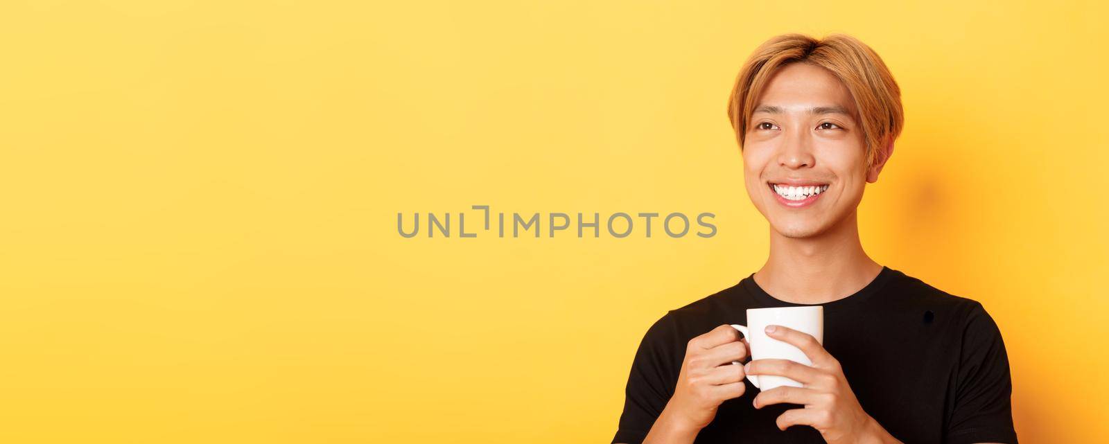 Close-up of happy smiling handsome blond asian guy, looking away with dreamy nostalgic look while drinking coffee, standing over yellow background by Benzoix