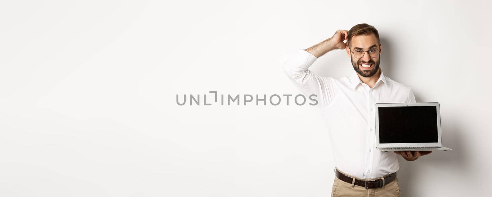 Awkward business man showing laptop screen and looking doubtful, standing against white background uncomfortable.