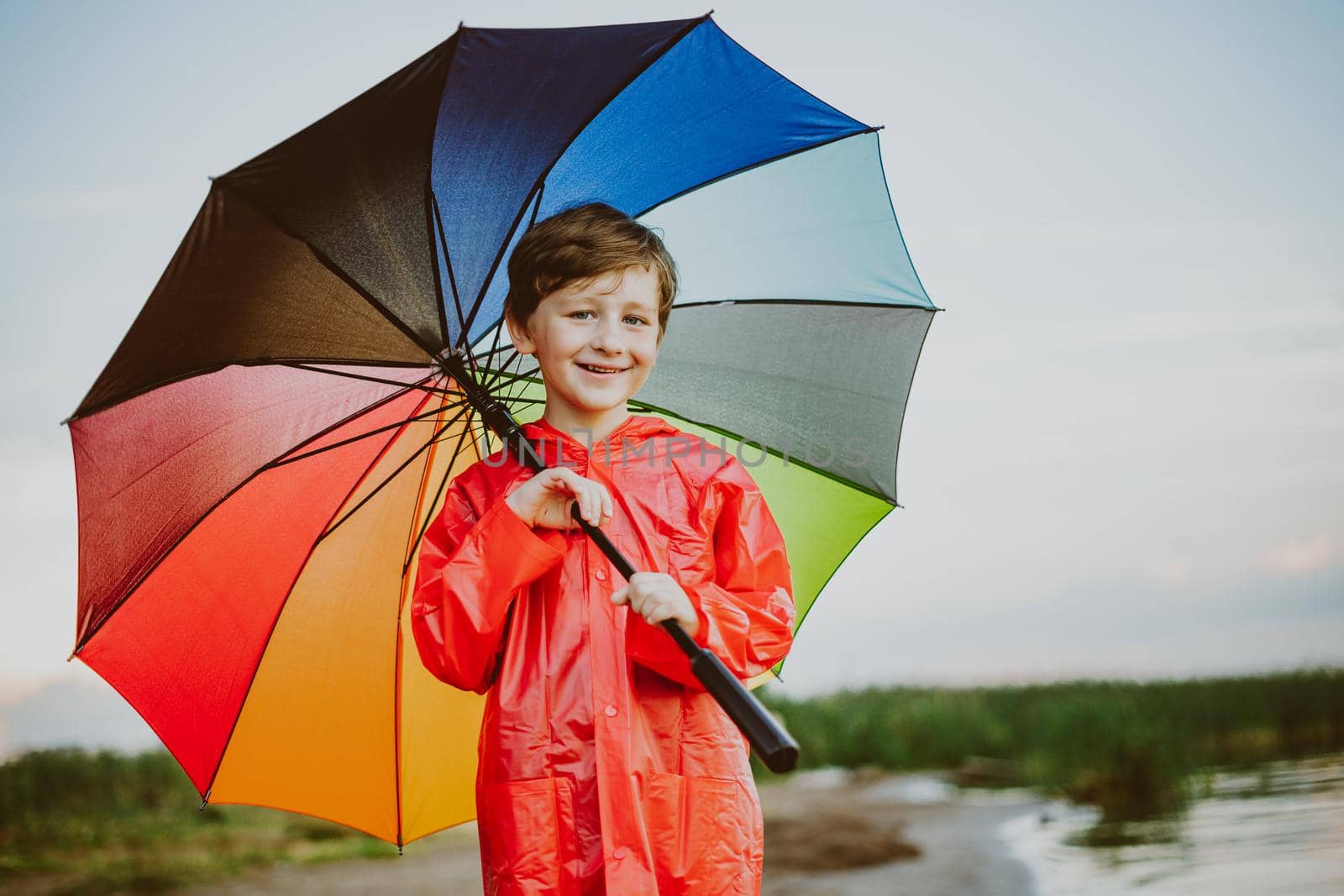 Portrait of a smiling school boy with rainbow umbrella in the park. Kid holds colourful umbrella on his shoulder. Cheerful child in a red raincoat holding multicolor umbrella outdoors.