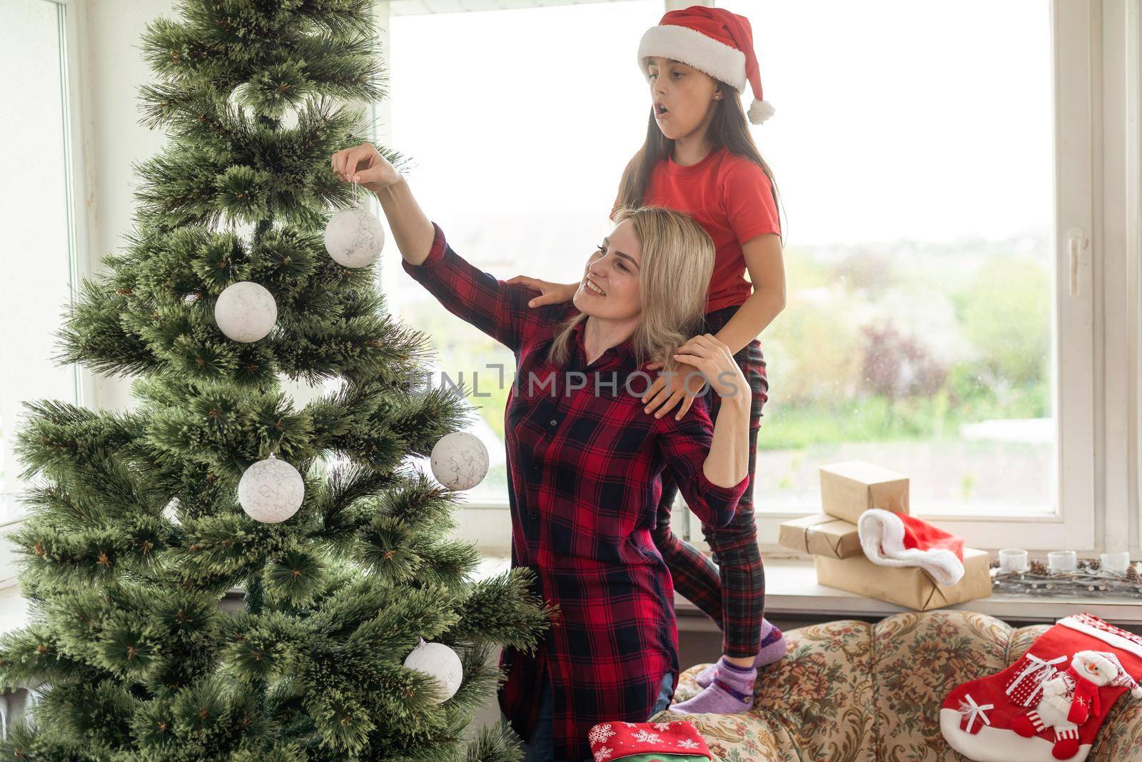 Attractive mother and daughter wearing winter outfits and decorating Christmas tree at home with joy, preparing to celebrate Christmas festival. Concept for family Christmas party