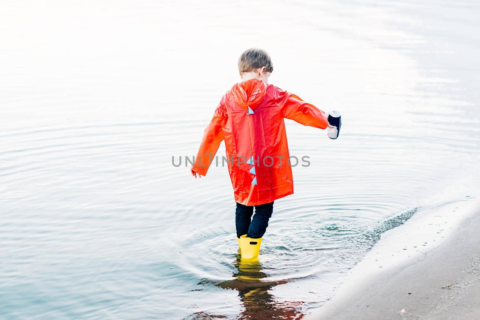 Boy in a red raincoat and yellow rubber boots playing with water at the beach. School kid in a waterproof coat jumping in water at sea. Child having fun with waves at the shore.