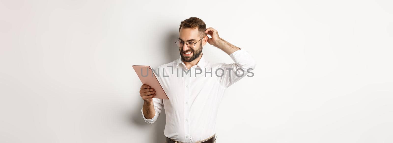 Confused male manager looking puzzled at digital tablet, scratching head doubtful, standing over white background.