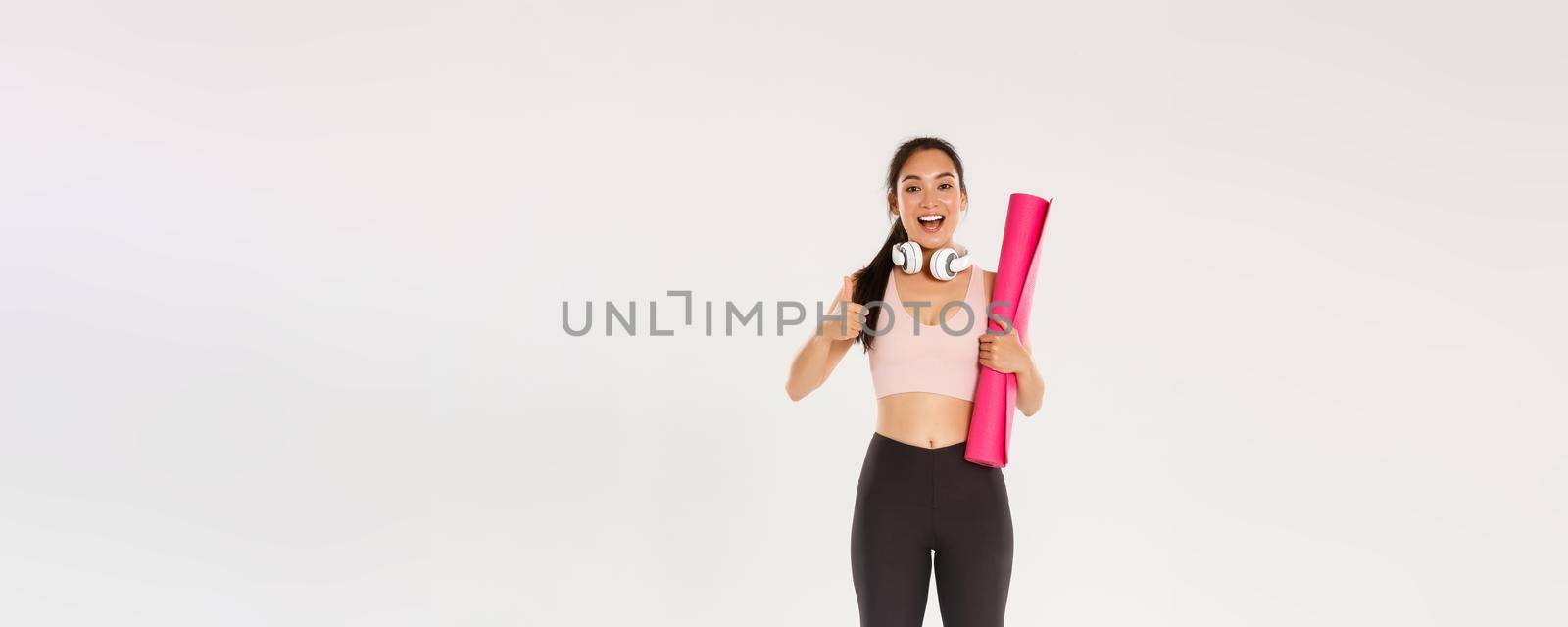 Full length of cheerful satisfied, slim asian girl with fitness rubber mat and headphones, female athlete showing thumbs-up, recommend gym or yoga classes, looking pleased after good workout.
