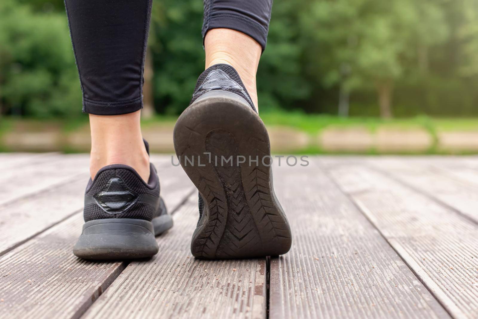 Close-up of women's legs in black sneakers and black leggings walking on a wooden deck in a park, in summer. Copy space