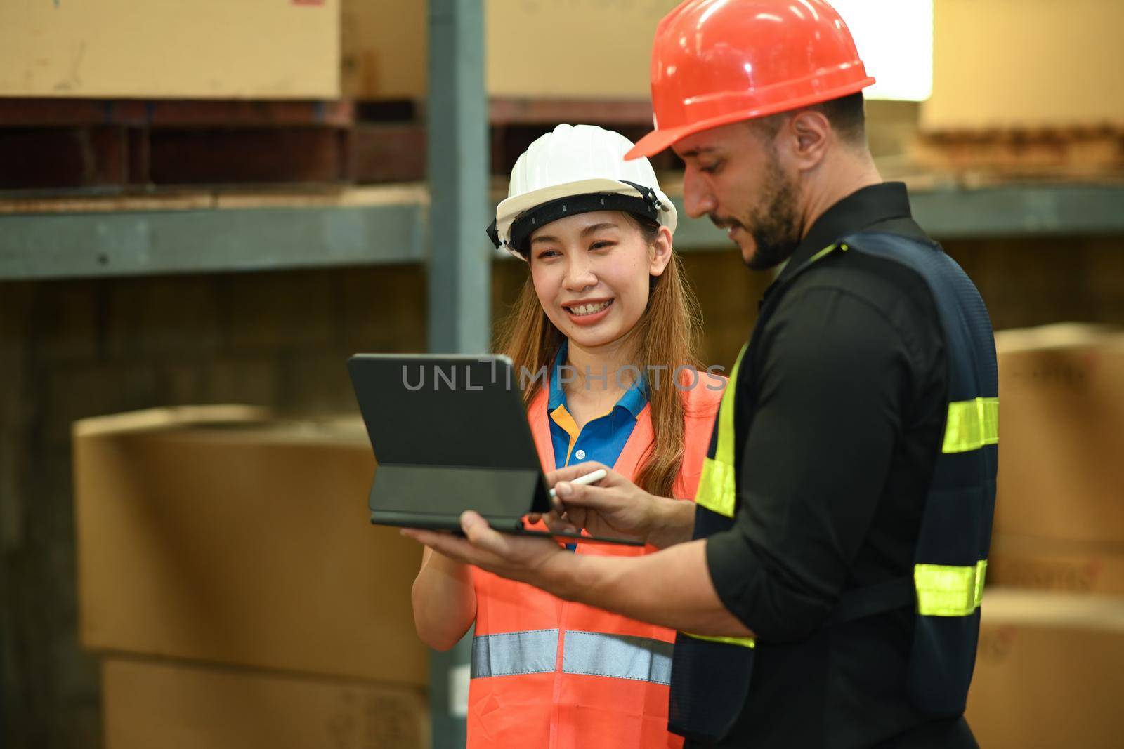Male manager and warehouse worker inspecting stock tick and cardboard stock product on digital tablet in a large warehouse.