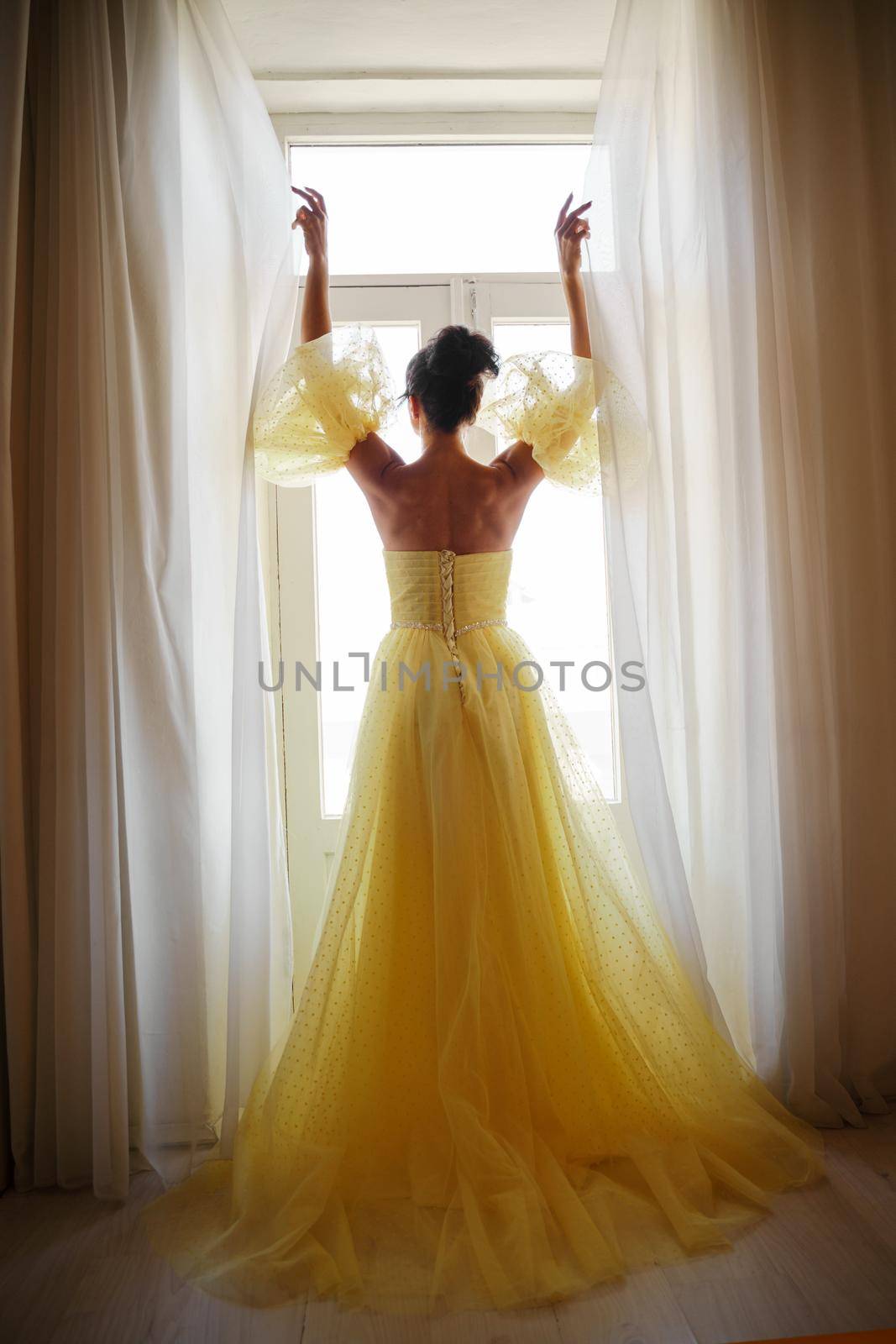 A woman's silhouette in a golden luxurious dress against the background of a window holds a curtain with her hands. Elegant lady in a yellow long silk dress with bare back, back view