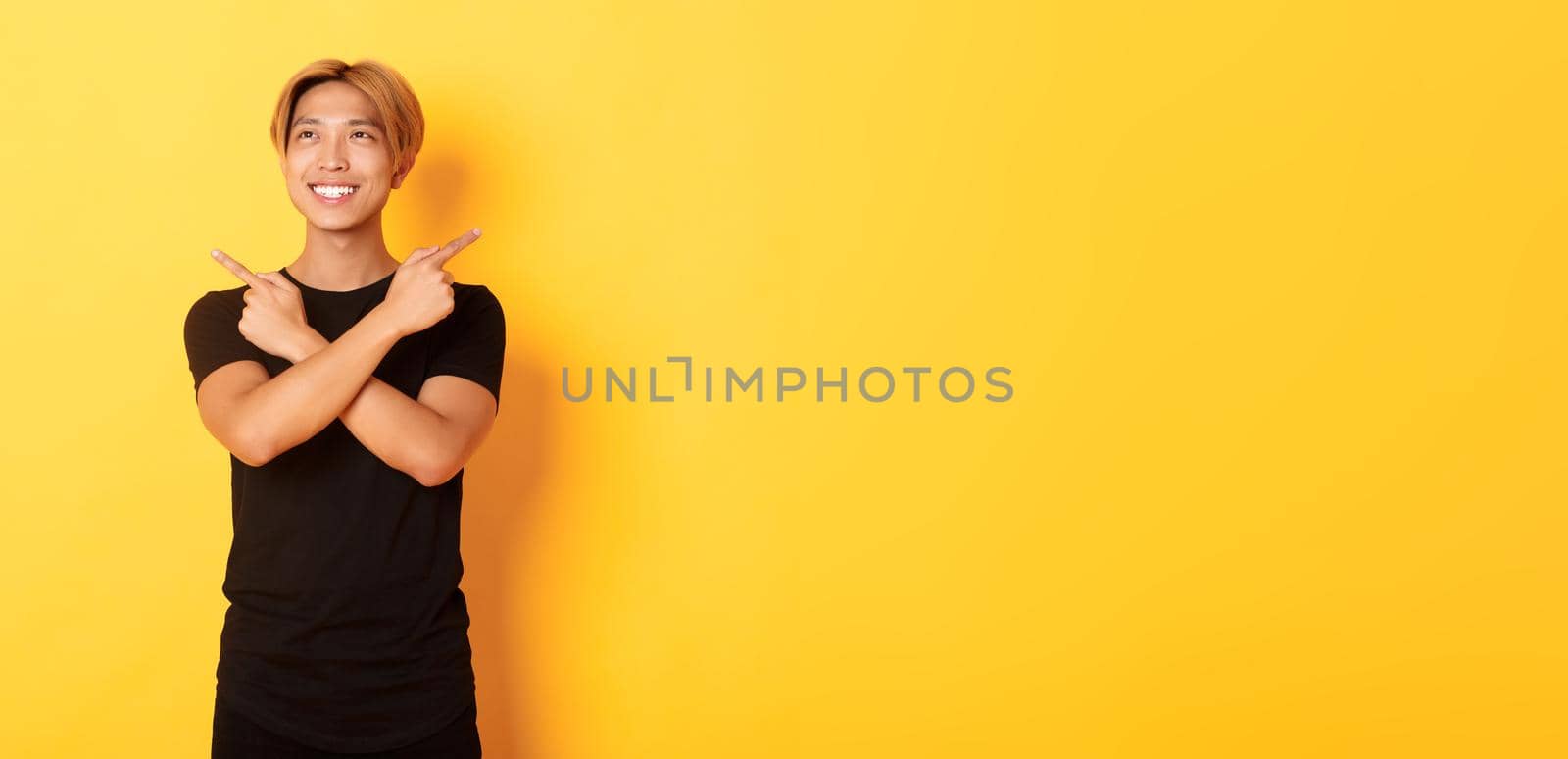 Portrait of smiling pleased asian guy, looking left and making decision, pointing sideways, yellow background by Benzoix