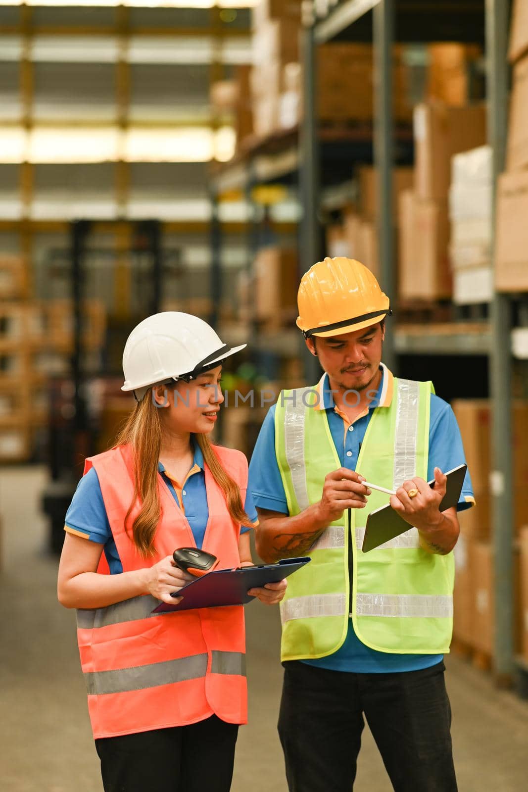 Warehouse workers in hardhats and and vests looking up order details on a tablet while standing between retail warehouse full of shelves.