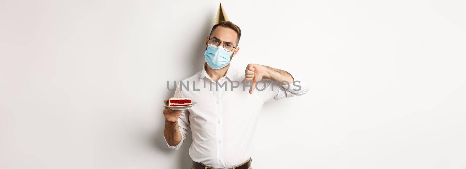Covid-19, social distancing and celebration. Disappointed birthday guy wearing face mask, holding bday cake with wish candle, making thumb down to express dislike, white background.
