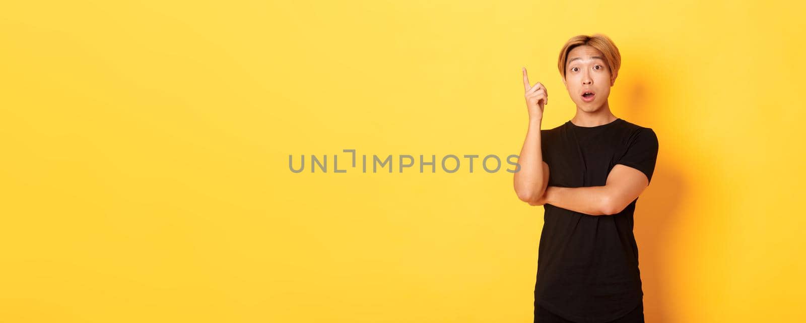 Portrait of thoughtful handsome asian man raising finger, suggesting idea, standing yellow background by Benzoix