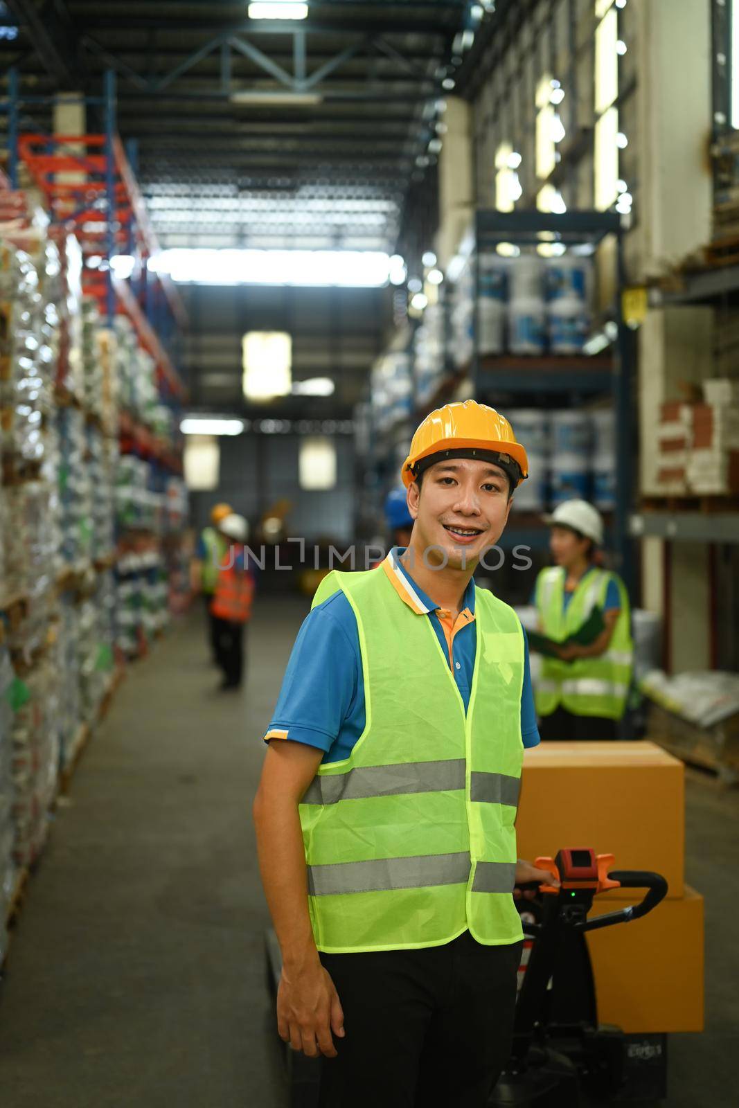 Male warehouse worker in hardhats and reflective jackets pulling a pallet truck walking through retail warehouse full of shelves.