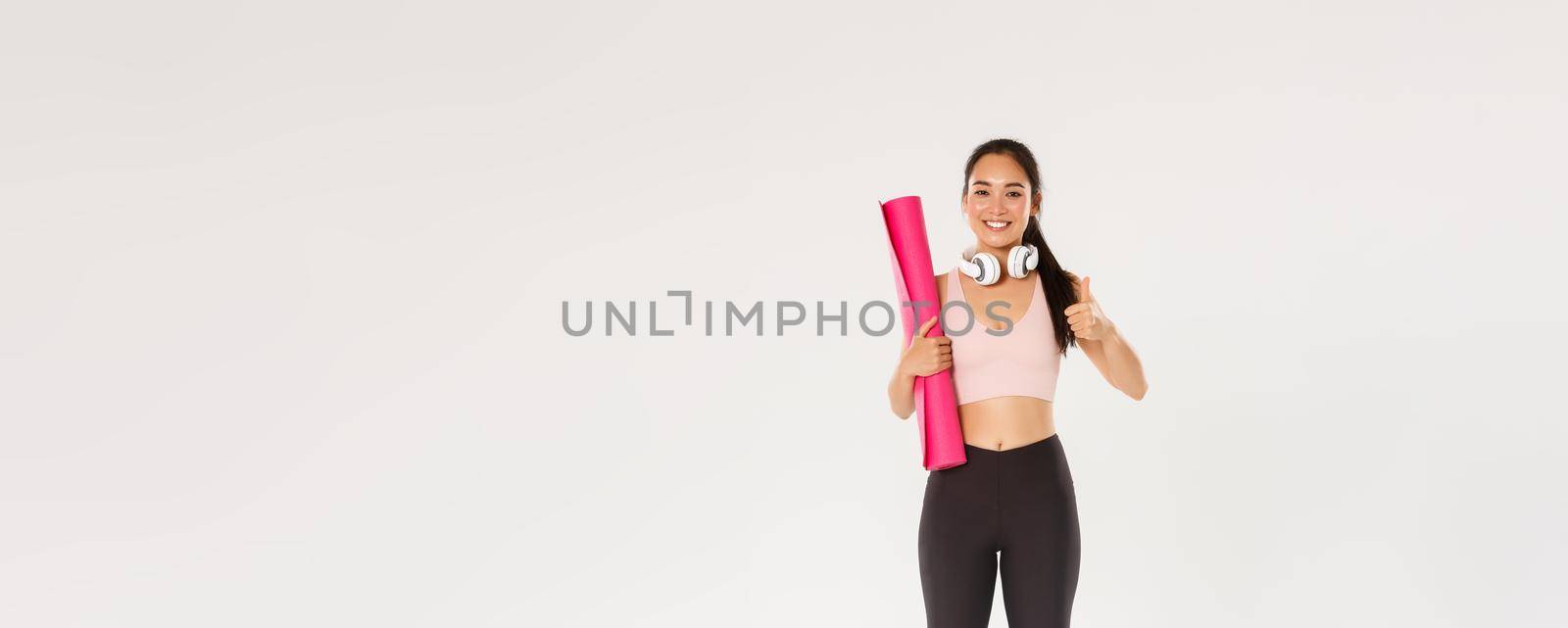 Full length of cheerful satisfied, slim asian girl with fitness rubber mat and headphones, female athlete showing thumbs-up, recommend gym or yoga classes, looking pleased after good workout.