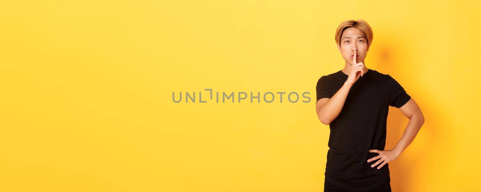 Portrait of attractive blond asian man shushing, press finger to lips and hush, yellow background.