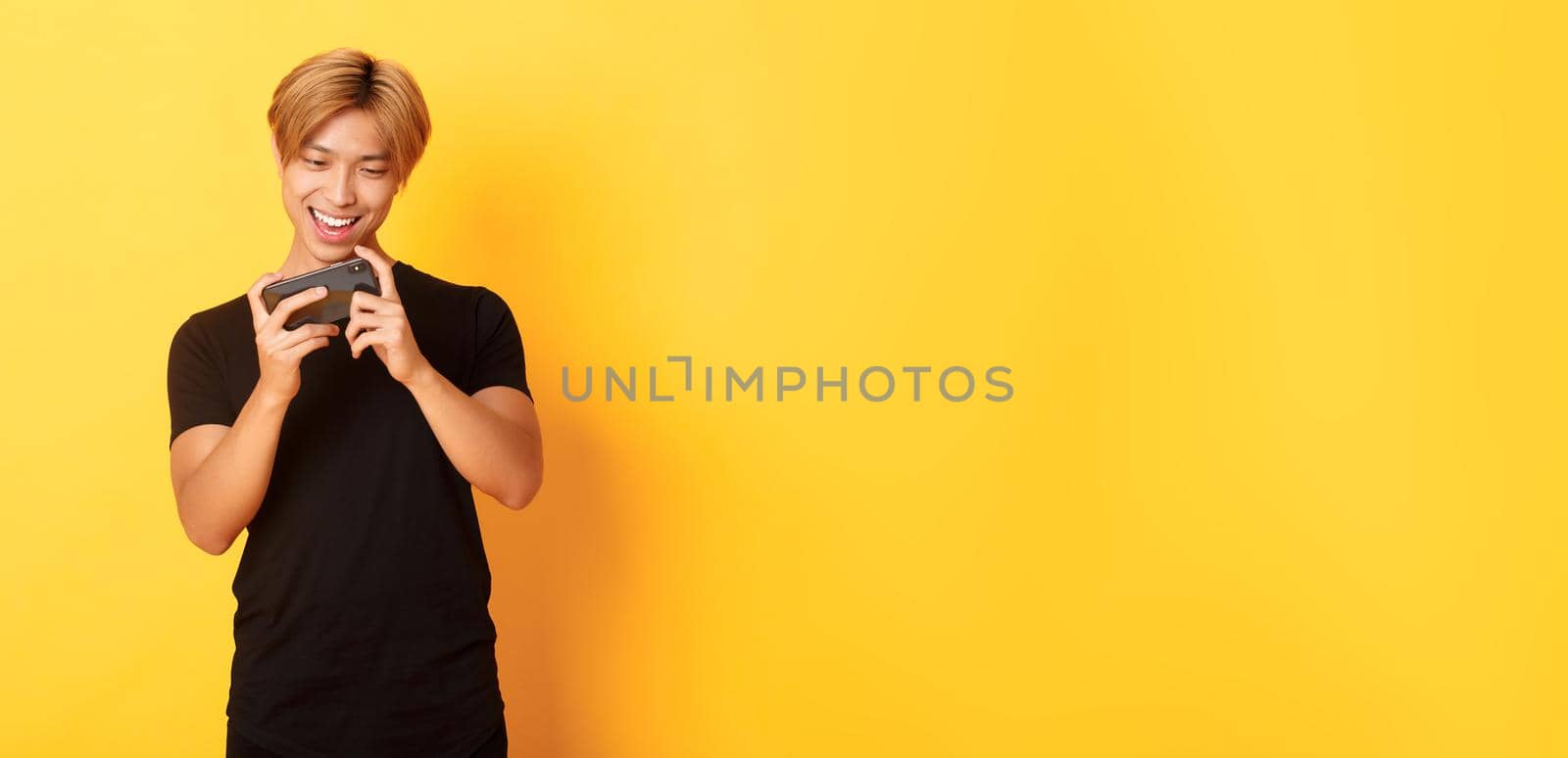 Portrait of stylish handsome asian guy playing smartphone game, looking amused at mobile screen, standing yellow background by Benzoix