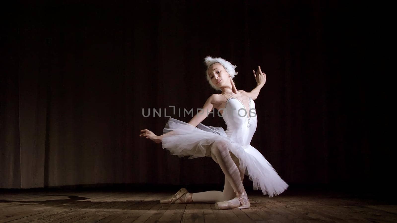 in rays of spotlight, on the stage of the old theater hall. Young ballerina in suit of white swan and pointe shoes, dances elegantly certain ballet motion, part de bras. High quality photo
