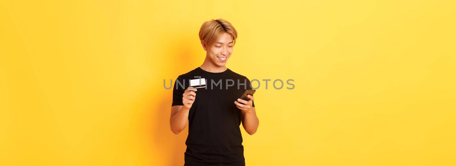 Smiling handsome asian man paying for online purchase, shopping in internet, holding smartphone and credit card, yellow background.