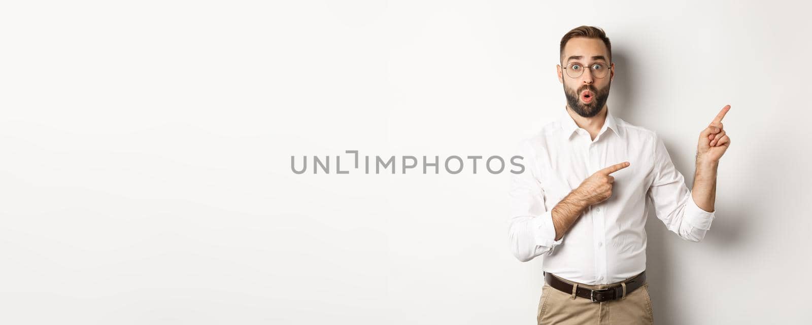 Impressed businessman showing interesting promo offer, pointing fingers right, standing amazed against white background.