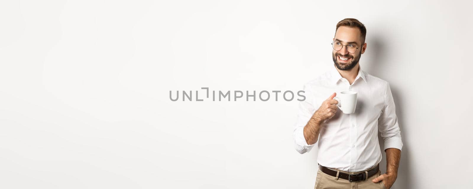Successful business man drinking coffee, looking sideways with satisfied smile, standing over white background.