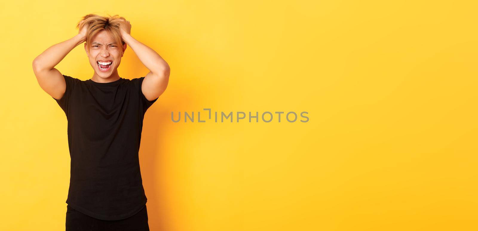 Portrait of pissed-off angry asian man tossing haircut and yelling furious, standing over yellow background.