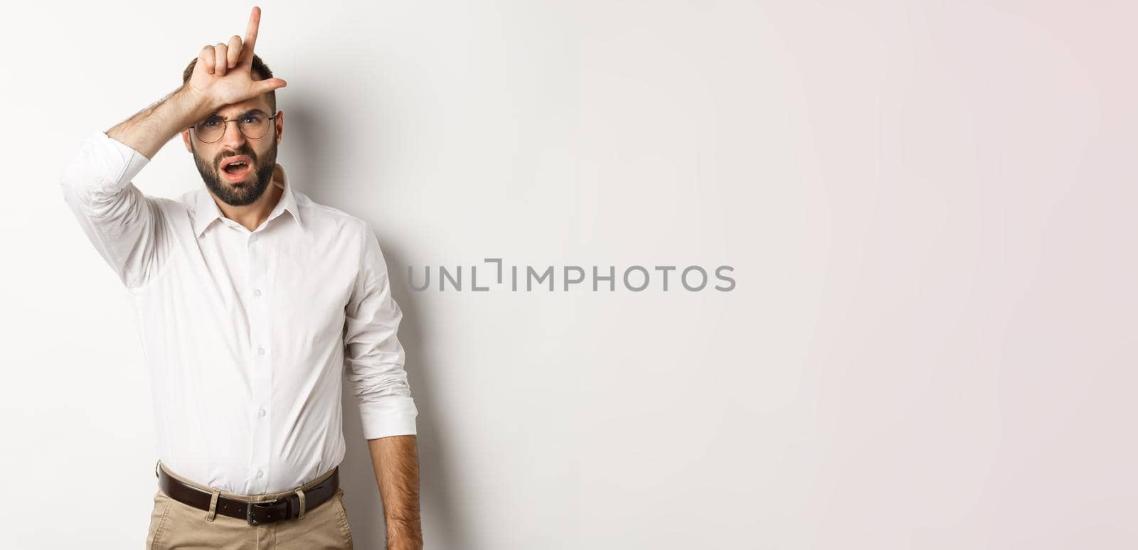 Shocked guy showing loser sign on forehead, complaining, standing over white background by Benzoix