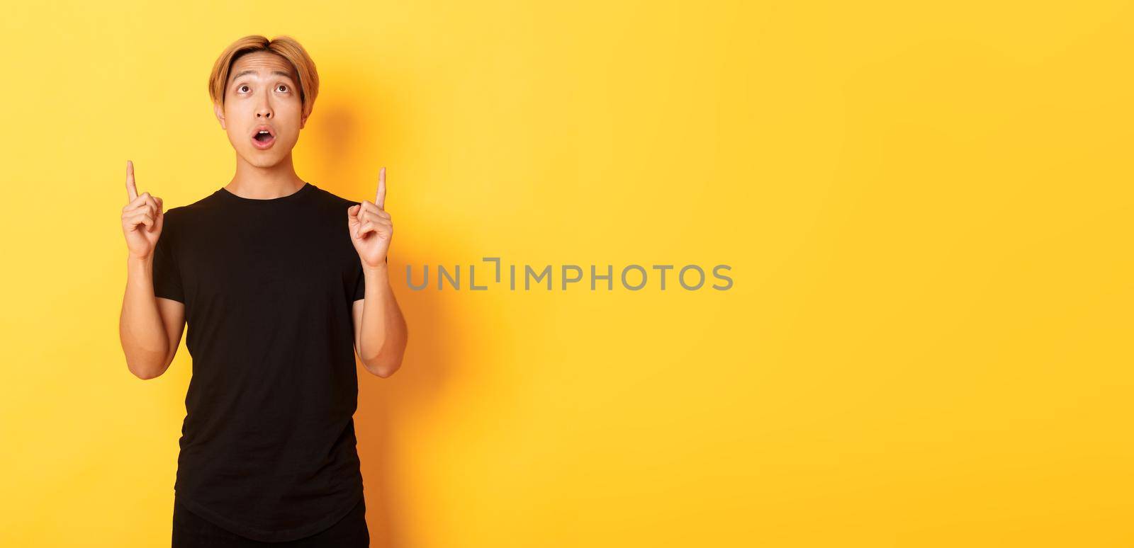 Portrait of curious and amazed asian guy with blond hair, wearing black t-shirt, looking and pointing fingers up astonished, yellow background.