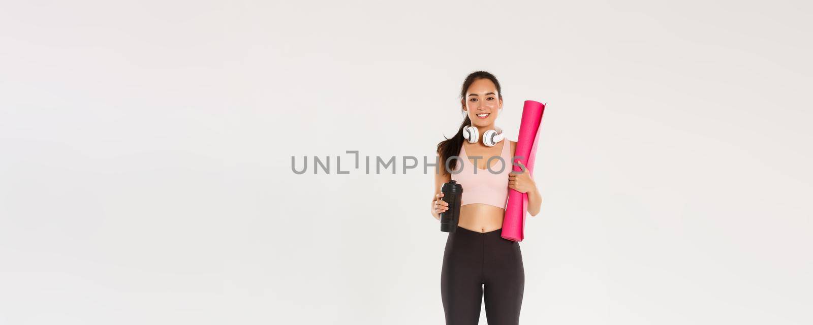 Full length of cute brunette asian fitness girl with headphones, activewear and water bottle, carry pink rubber mat for gym exercises, going for yoga classes, standing over white background by Benzoix