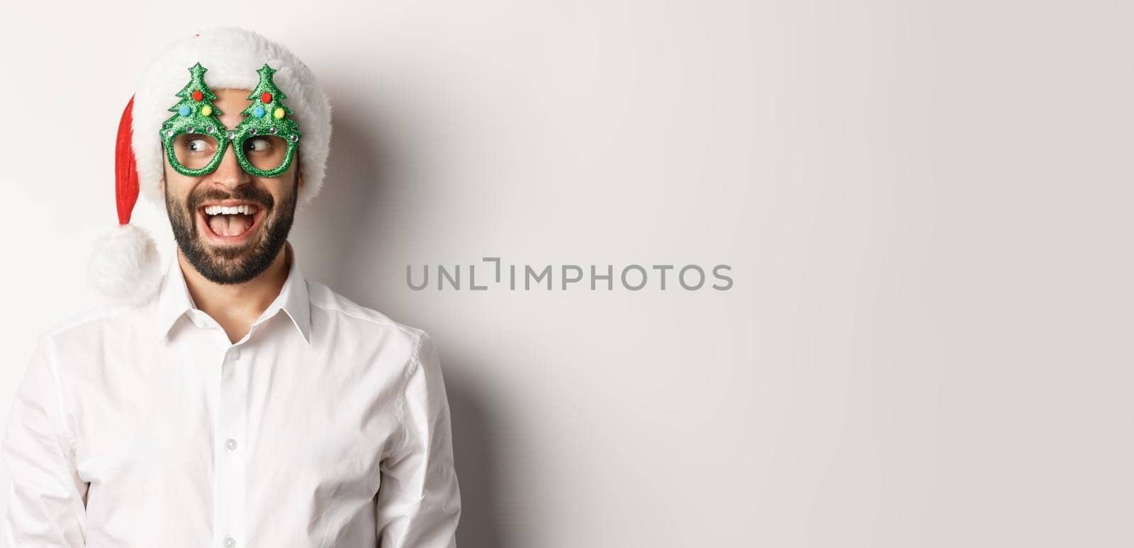 Close-up of funny man looking left with surprised face, wearing christmas party glasses and santa hat, celebrating New Year, white background.