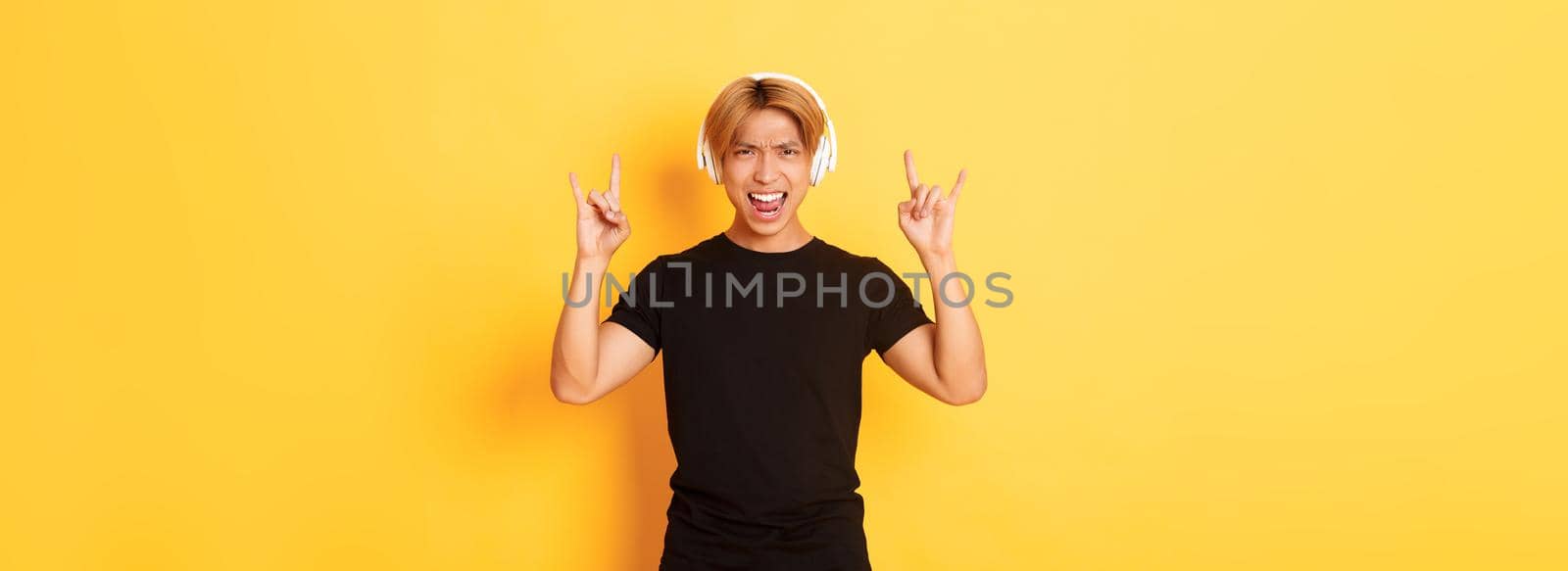 Joyful handsome asian guy enjoying listening music in headphones, showing rock-n-roll gesture, standing over yellow background.