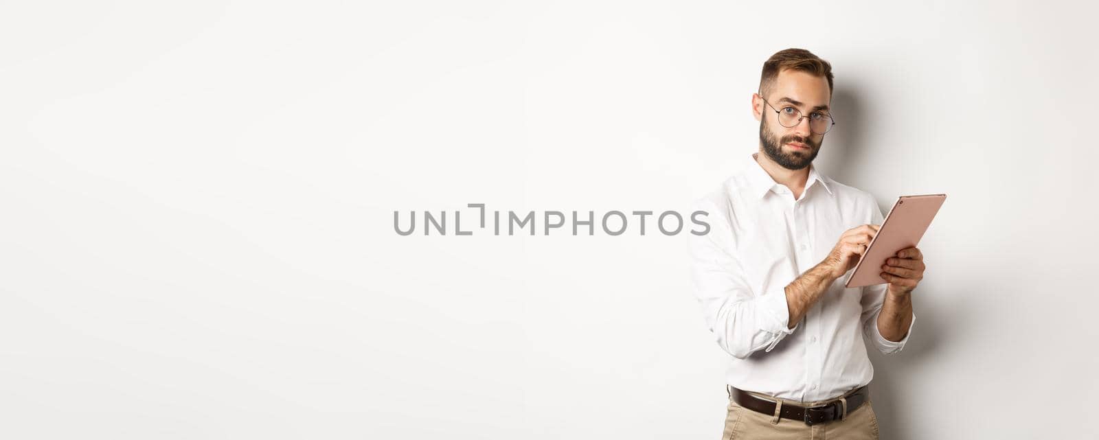 Handsome businessman doing job on digital tablet, reading something, standing over white background by Benzoix