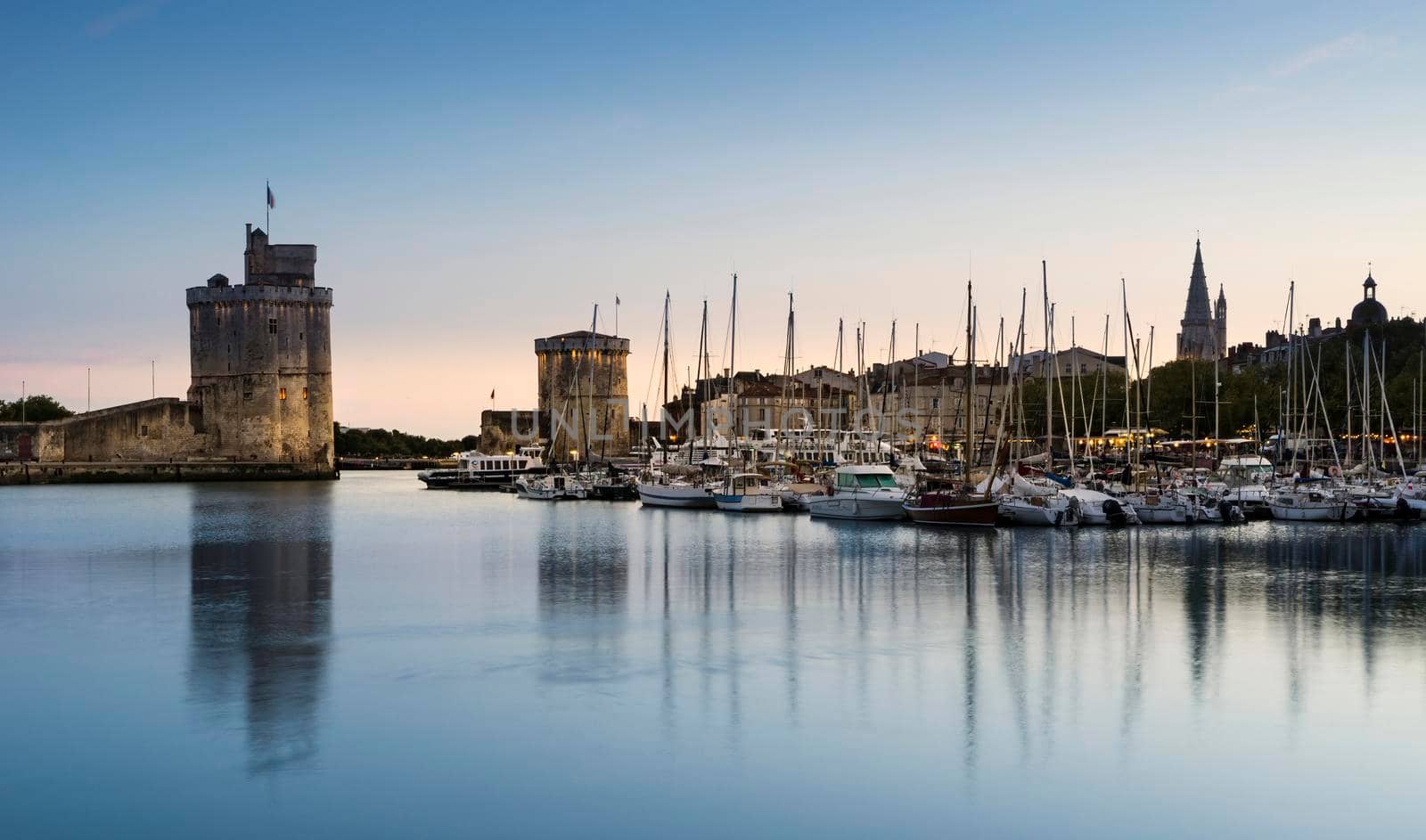 View of the old port of the French city of La Rochelle at sunset with ships at anchor in the harbour and the two towers in the background.