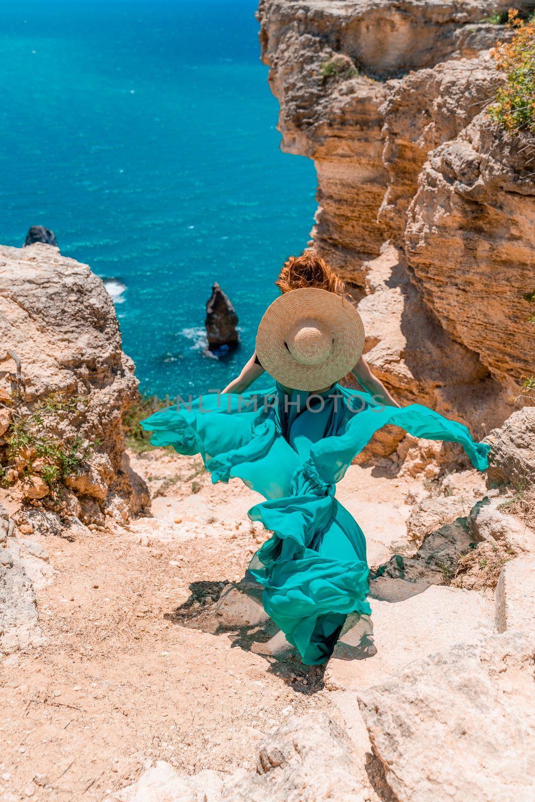 A girl with loose hair in a long mint dress descends the stairs between the yellow rocks overlooking the sea. A rock can be seen in the sea. Sunny path on the sea from the rising sun by Matiunina