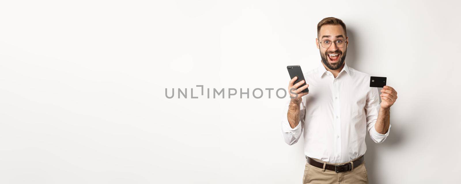 Business and online payment. Excited man paying with mobile phone and credit card, smiling amazed, standing over white background.