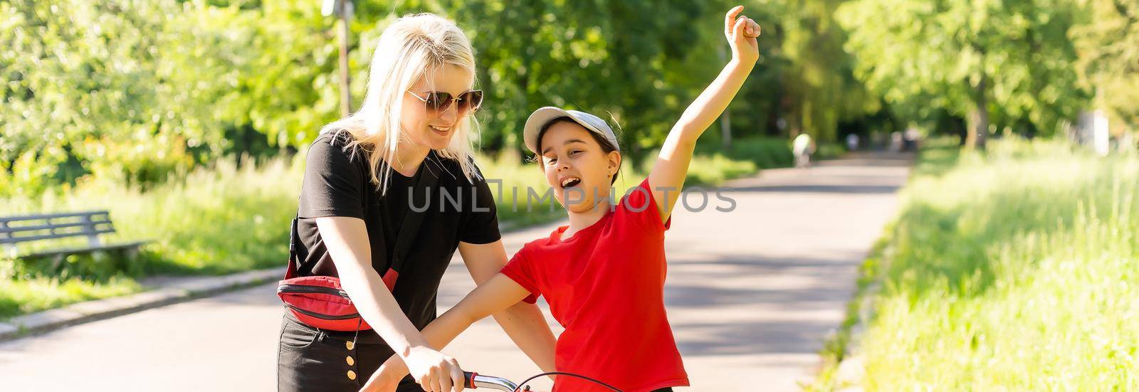 Beautiful and happy young mother teaching her daughter to ride a bicycle. Both smiling, summer park in background. by Andelov13