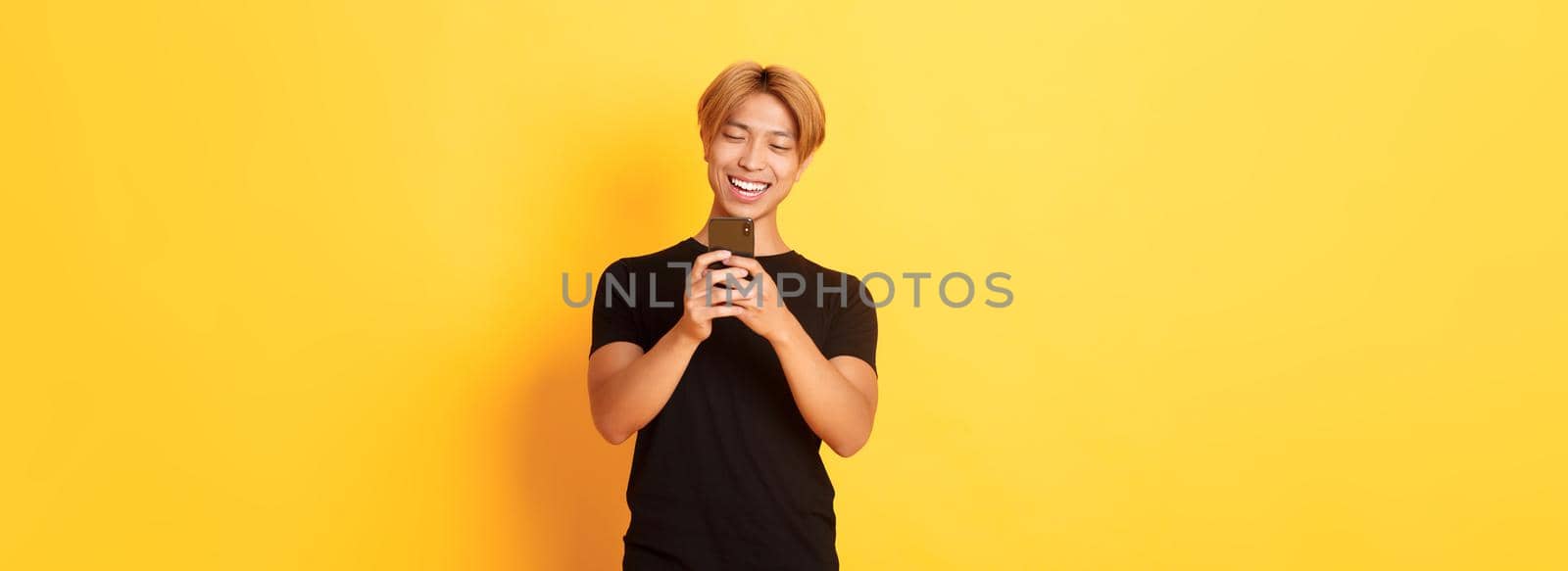 Portrait of handsome stylish asian guy with blond hair, using mobile phone and smiling, messaging in smartphone app, yellow background.
