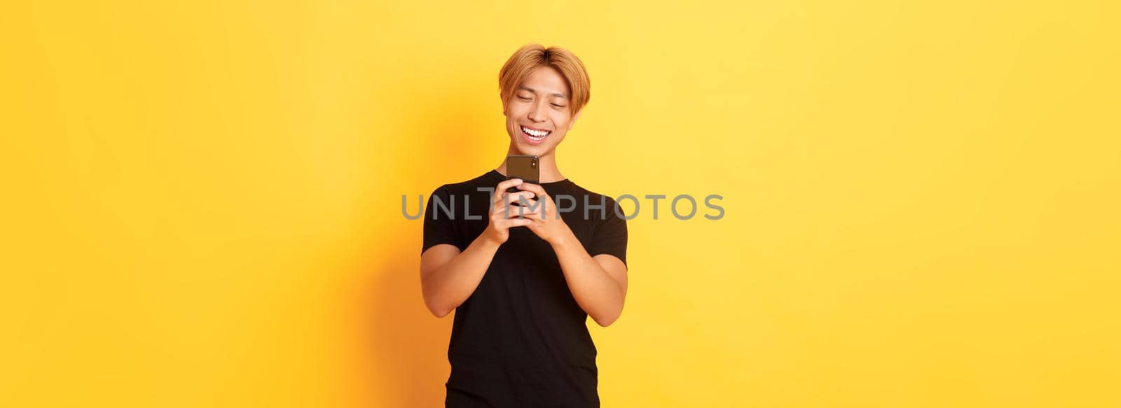 Portrait of handsome stylish asian guy with blond hair, using mobile phone and smiling, messaging in smartphone app, yellow background by Benzoix